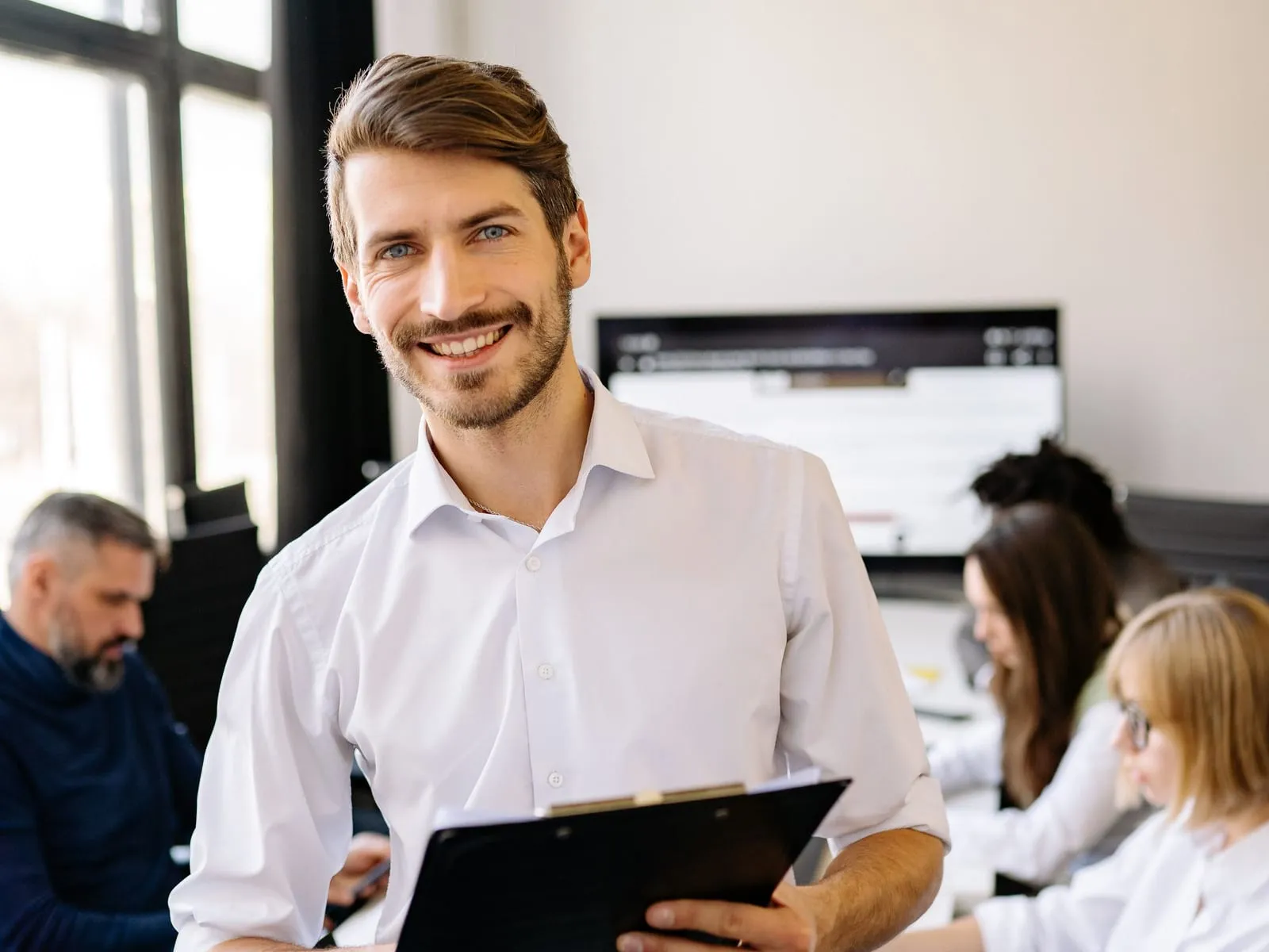 A smiling professional faces the camera while colleagues work in a bright office.